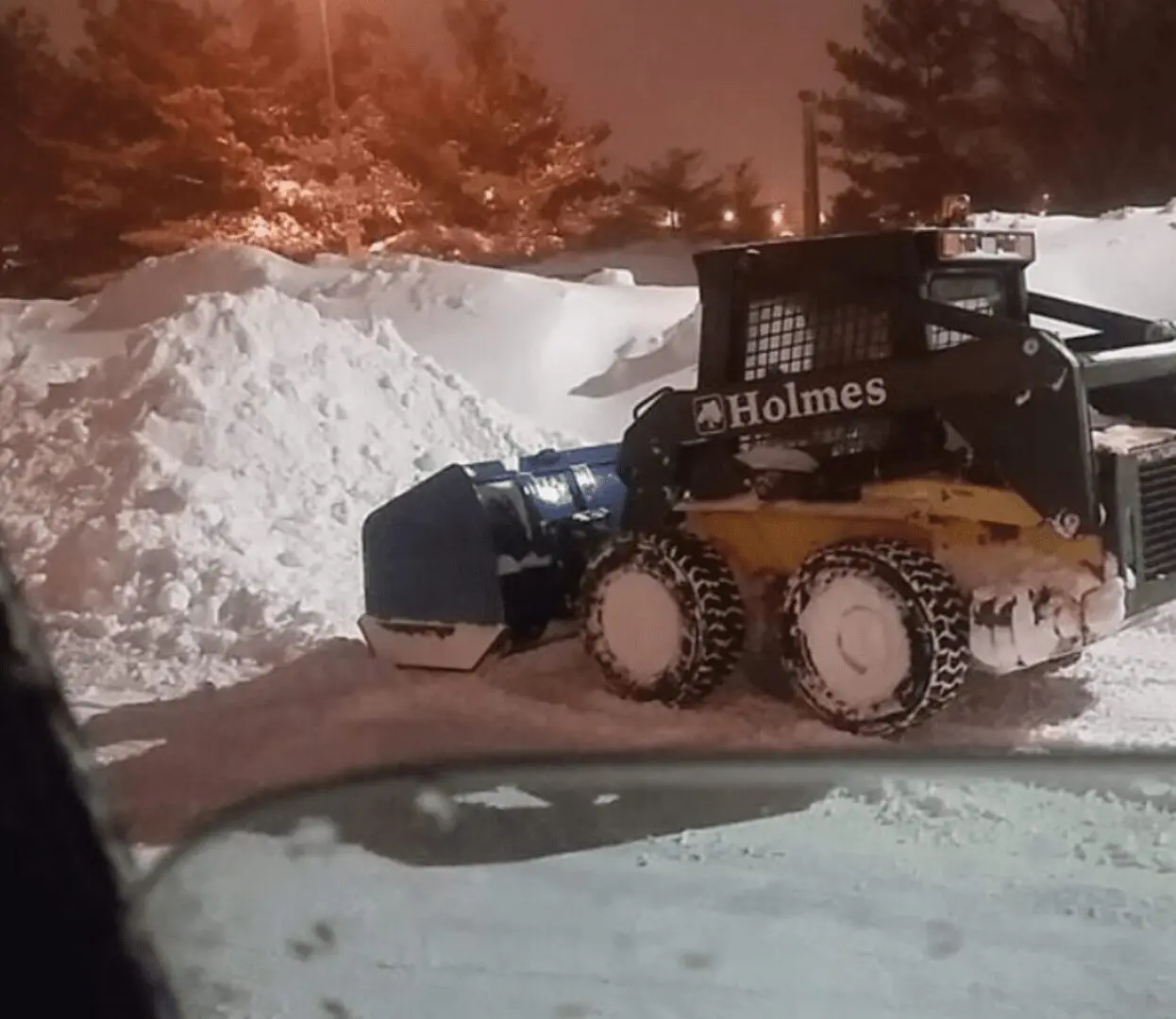 A small machine clears snow on a driveway at night.